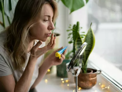 Woman applying moisturizing nourishing balm to her lips with her finger to prevent dryness and chapping in the cold season, sitting by the window, looking at mirror. Lip protection.