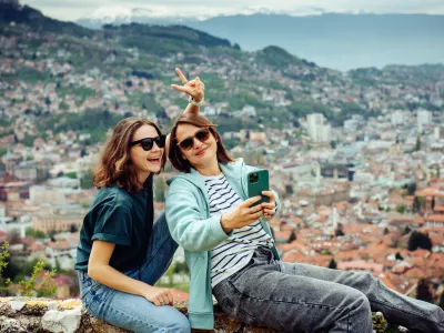 Two cheerful traveler girls in dark glasses are having fun and taking pictures against the background of Sarajevo, a beautiful European city in the mountains.