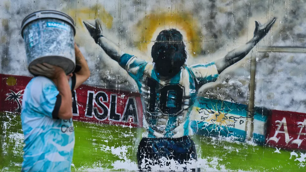 A pedestrian carries a bucket past graffiti depicting late soccer legend Diego Maradona in Buenos Aires, Argentina, Monday, April 13, 2026. (AP Photo/Rodrigo Abd)