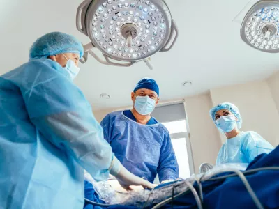 Low angle portrait of experienced male surgical and team making operation in sterile medical room. Two female assistants help him. Minimally invasive surgery. / Foto: Iryna Inshyna