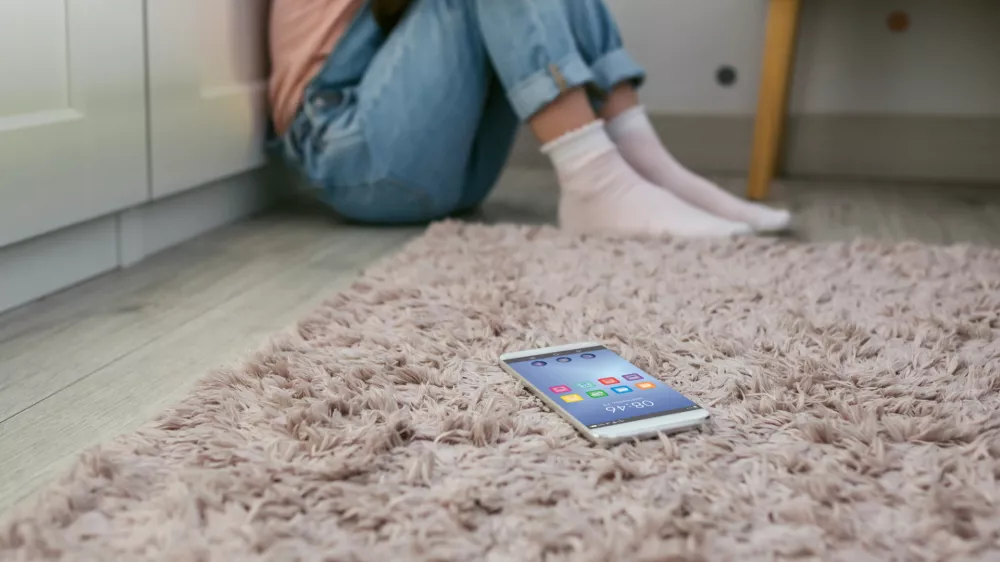 Mobile phone lying on the floor with unrecognizable sad little girl sitting behind. Selective focus on mobile in foreground