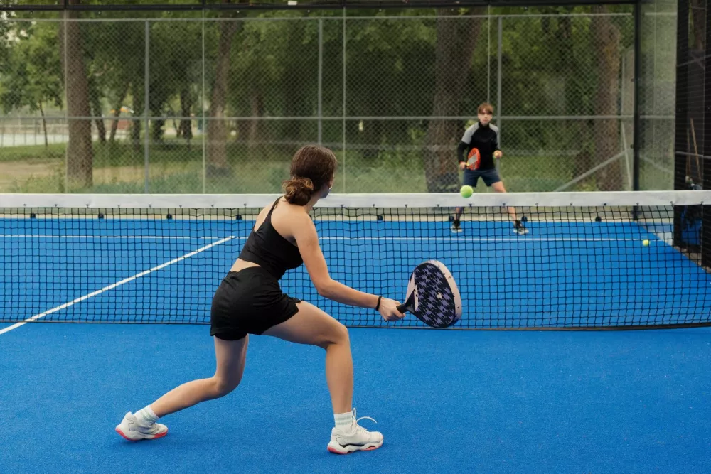 Two young players engaging in a competitive game of padel tennis on a blue outdoor court surrounded by a green park. / Foto: Arthurhidden
