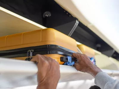 A person's hands are seen placing or retrieving a yellow hard-shell suitcase from an airplane's overhead storage compartment.