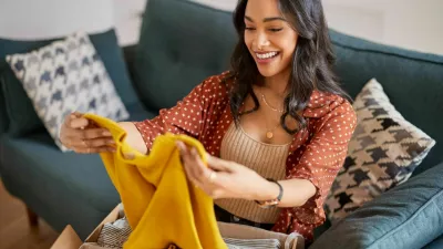 Happy young latin woman checking clothes sitting on sofa at home. Cheerful shopaholic hispanic woman holding new clothes. Smiling multuethnic girl unpacking parcel box with clothes after shopping online.